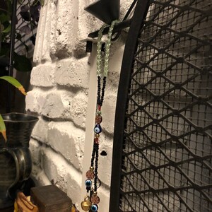May include: A black beaded necklace with blue evil eye charms and gold bells hanging from it. The necklace is hanging on a black metal grate against a white brick wall.