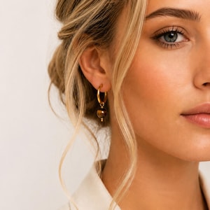 May include: Gold hoop earrings with a small, round, brown stone pendant. The earrings are worn on a person with blonde hair and a cream-colored top. The earrings are a simple, elegant design.