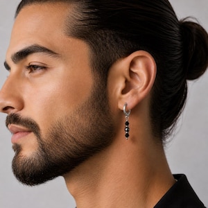 May include: A silver hoop earring with three black beads hanging from it. The earring is worn on the left ear. The person in the image has dark hair pulled back in a bun and a beard.