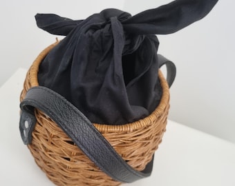 May include: A round, woven brown wicker basket bag featuring a black fabric drawstring closure tied at the top. The bag includes a black strap attached to the sides with silver-toned metal rivets, providing a functional handle for carrying.