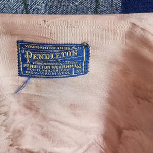 以下が含まれることがあります： 「WARRANTED TO BE A」と「PENDLETON」の文字が入った、青と金のペンドルトンラベルのクローズアップ。ラベルには「TRADE MARK REG. U.S. PAT. OFF」、「PENDLETON WOOLEN MILLS PORTLAND, OREGON」と「100% VIRGIN WOOL」の文字も含まれています。ラベルはピンク色の生地にあります。