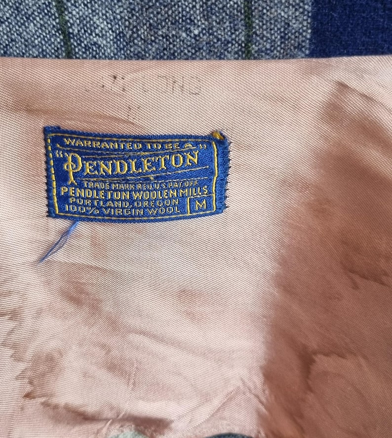 以下が含まれることがあります： 「WARRANTED TO BE A」と「PENDLETON」の文字が入った、青と金のペンドルトンラベルのクローズアップ。ラベルには「TRADE MARK REG. U.S. PAT. OFF」、「PENDLETON WOOLEN MILLS PORTLAND, OREGON」と「100% VIRGIN WOOL」の文字も含まれています。ラベルはピンク色の生地にあります。