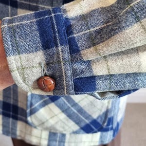 以下が含まれることがあります： 青、白、グレーのチェック柄ジャケットの袖のクローズアップ。袖には茶色の革ボタンとポケットが付いています。ジャケットはチェック柄の衣装の一部です。