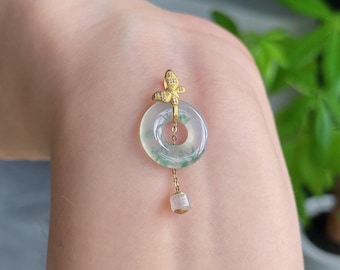 Op de afbeelding: Een gouden hanger met een cirkelvormige, lichtgroene jade donut met een vlinderaccent aan de bovenkant. De vlinder is versierd met kleine heldere steentjes. Een dunne gouden ketting hangt in het midden van de jade en eindigt in een kleine cilindrische kraal.