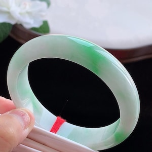 Könnte beinhalten: Ein glatter, polierter Jade-Armreif mit einer durchscheinenden weißen Basis und lebhaften, gesprenkelten grünen Flecken auf dem gesamten kreisförmigen Band. Der Stein hat eine glänzende, reflektierende Oberfläche, die das Licht einfängt.