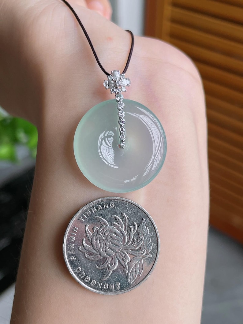 Op de afbeelding: Een lichtgroene jade hanger met een zilveren en diamant accent, hangend aan een zwart koord. De hanger is rond en glad. Een zilveren munt met een bloemenontwerp is ook zichtbaar.
