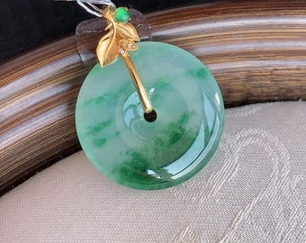 Op de afbeelding: Een cirkelvormige groene jade hanger met een gepolijste afwerking en natuurlijke gevlekte patronen. De hanger is voorzien van een goudkleurig metalen oogje in de vorm van een takje met blaadjes, geaccentueerd door een klein groen kraaltje en kleine heldere steentjes.