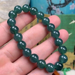 May include: A dark green beaded bracelet. The bracelet is made of round, polished beads. The beads have a glossy finish and reflect light. The bracelet is held in a hand. The bracelet is a piece of jewelry.