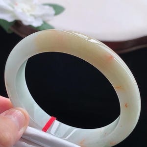 May include: A smooth, polished jade bangle bracelet featuring a translucent pale green base with a prominent warm honey-colored section and small, scattered reddish-orange inclusions throughout the circular stone band.