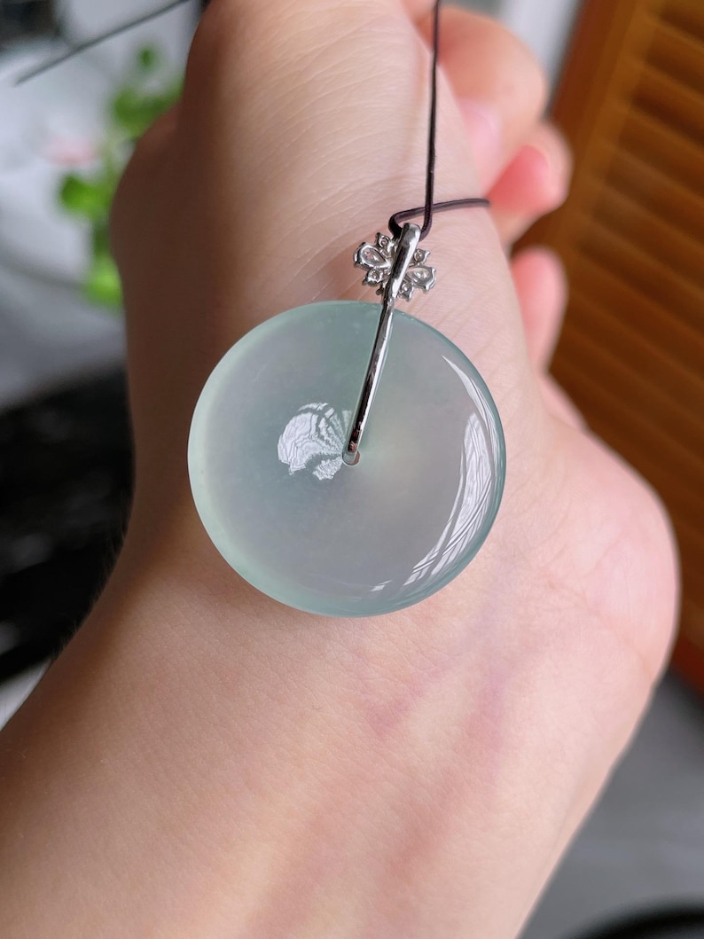 Op de afbeelding: Een ronde, lichtgroene jade hanger met een zilverkleurige balein en een zwart koord. De hanger is glad en doorschijnend, met een subtiel, natuurlijk patroon. De balein is versierd met een bloemmotief. Sieraden.