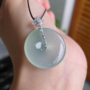 Op de afbeelding: Een ronde, lichtgroene jade hanger met een diamantaccent en een zwart koord. De jade heeft een glad, gepolijst oppervlak. Het diamantaccent is een cluster van kleine stenen die leiden naar een rij kleinere diamanten.