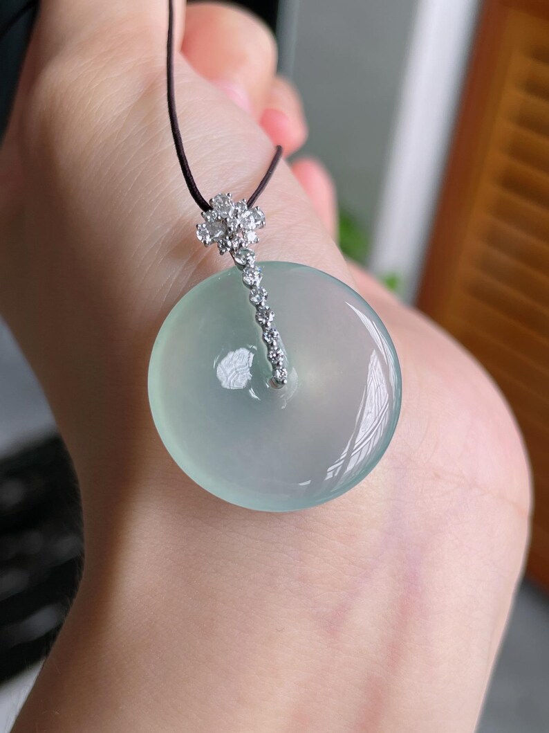 Op de afbeelding: Een ronde, lichtgroene jade hanger met een diamantaccent en een zwart koord. De jade heeft een glad, gepolijst oppervlak. Het diamantaccent is een cluster van kleine stenen die leiden naar een rij kleinere diamanten.