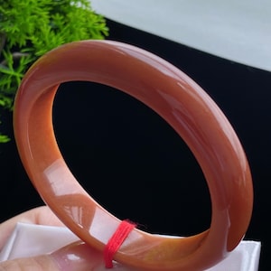 Puede incluir: Una pulsera tipo brazalete de color naranja rojizo, lisa y pulida, con un perfil redondeado. La pulsera presenta un acabado brillante y translúcido, con un pequeño cordel rojo fino atado alrededor de una sección del aro, resaltando su forma.