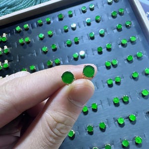 May include: A pair of round, vivid green jade stud earrings with gold settings. The earrings are held in a hand, with many other similar earrings visible in the background, displayed on a black tray.