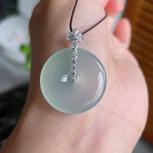 Op de afbeelding: Een ronde, lichtgroene jade hanger met een zilveren en diamant accent. De hanger hangt aan een dun, donkerbruin koord. Het zilveren accent heeft een bloemenmotief aan de bovenkant en een rij diamanten. De jade heeft een doorschijnend uiterlijk.