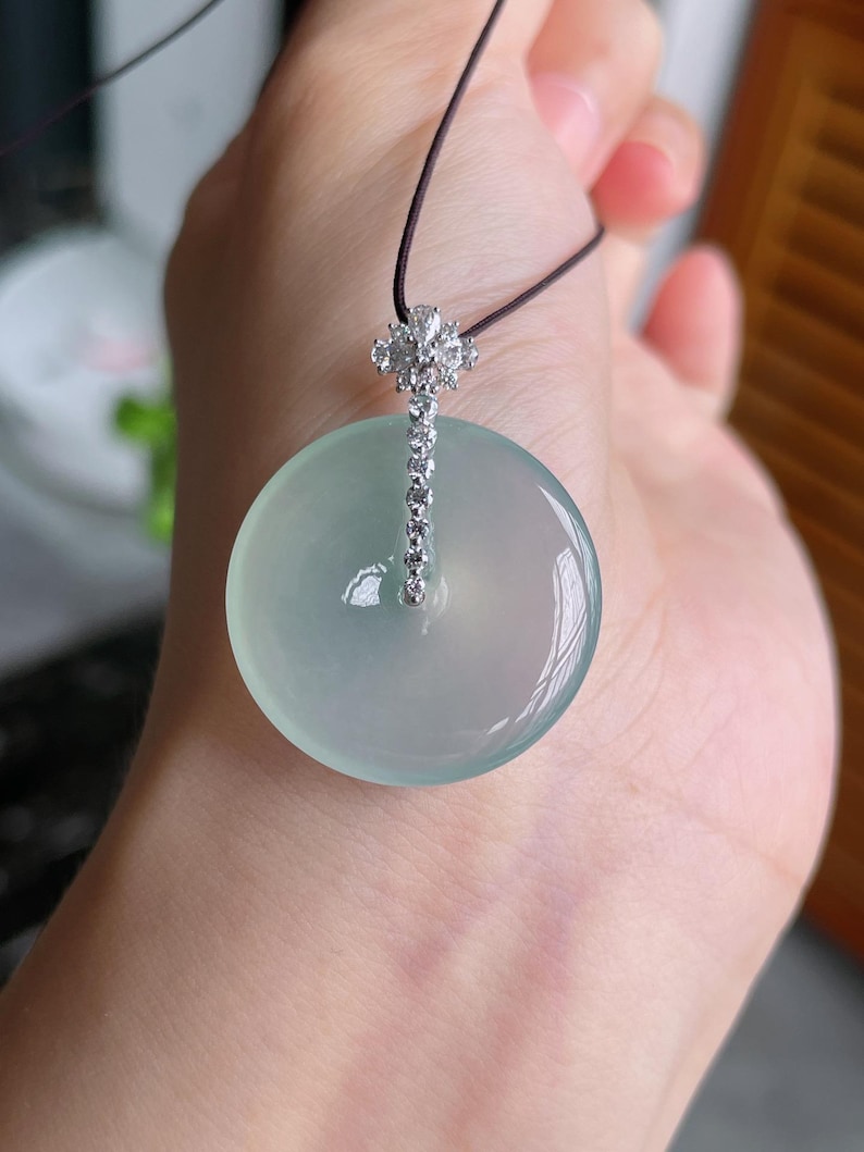 Op de afbeelding: Een lichtgroene jade hanger met een zilveren en diamant accent. De hanger is rond en heeft een glad oppervlak. Het zilveren accent is een verticale staaf met diamanten en een bloemmotief aan de bovenkant. Hij hangt aan een zwart koord.