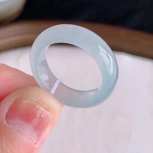 May include: A smooth, polished band ring crafted from translucent, pale icy-blue jadeite stone. The ring features a rounded, continuous circular profile with a glossy, reflective surface that catches the light, showcasing its cool, uniform color.