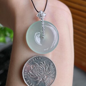 Op de afbeelding: Een ronde, lichtgroene jade hanger met een met diamanten bezette zilveren ketting en een zwart koord. De hanger wordt getoond naast een zilveren munt met een bloemmotief.