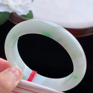 Könnte beinhalten: Ein glatter, polierter Jade-Armreif mit einer durchscheinenden weißen Basis und subtilen, leuchtend grünen Einschlüssen im gesamten kreisförmigen Band. Ein dünner roter Faden ist um den unteren Teil des polierten Schmuckstücks aus Stein gebunden.