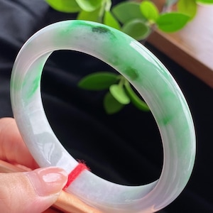 May include: A smooth, circular bangle bracelet crafted from polished jadeite stone. The bangle features a translucent white base color with vibrant, natural green inclusions and patches distributed throughout the rounded, solid stone band.