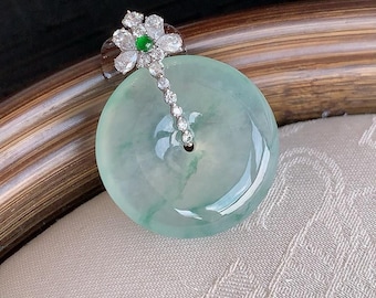 Op de afbeelding: Een ronde, lichtgroene jade hanger met een gepolijst, doorschijnend oppervlak. De hanger is voorzien van een zilverkleurige metalen hanger in de vorm van een bloem, bezet met heldere witte stenen en een klein groen steentje in het midden.