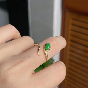 May include: A gold-colored ring in the shape of a snake, with a green gemstone at the head. The ring has a curved design, with small clear stones along the band. The snake's tail wraps around the finger.