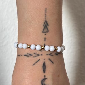 Peut inclure: Bracelet de perles blanches avec des accents dorés. Le bracelet est composé de perles blanches rondes alternées avec de petites perles dorées. Le bracelet est porté sur un poignet tatoué.