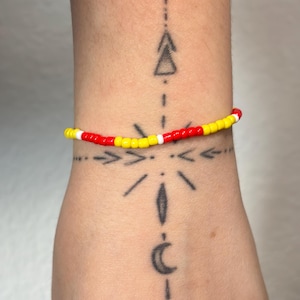 Peut inclure: Un bracelet de perles avec des perles rouges, jaunes et blanches. Le bracelet est porté sur un poignet avec un tatouage de formes et de lignes géométriques. Le bracelet est un accessoire coloré.