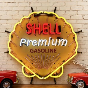 Puede incluir: Un letrero de neón con forma de concha que presenta un borde amarillo, el texto rojo SHELL, el texto blanco Premium y el texto rojo oscuro GASOLINE sobre un fondo dorado. El letrero está montado con soportes negros en una pared de ladrillo blanco.