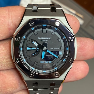 May include: A G-Shock Casio watch with a metallic silver and black octagonal bezel, featuring a dark gray dial with bright blue accents on the hands and hour markers. The watch includes a digital display window, analog hands, and a metal link band.
