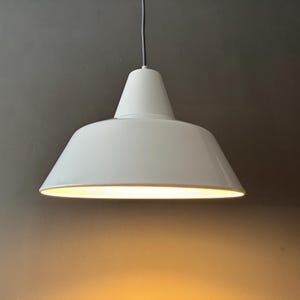 May include: A white, cone-shaped pendant light fixture with a wide, downward-facing shade. The light is suspended from a silver cord and illuminates a warm, yellow glow. The fixture is a modern design.