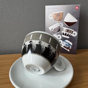以下が含まれることがあります： 白の陶器製エスプレッソカップとソーサーのセット。黒と白のフィルムストリップのグラフィックと人物のシルエットが描かれています。カップの底には「illy collection 2001」「10528」「made in Italy by IPA」の文字があります。プロモーションカードには他のカップのデザインが示されています。