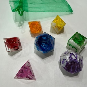 May include: A set of six multi-colored polyhedral dice featuring glittery resin interiors and dark numbers, accompanied by a sheer green drawstring pouch. The collection includes red, orange, yellow, green, blue, and purple translucent dice.