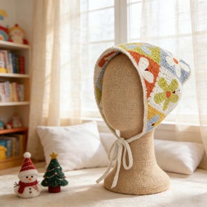 May include: A crochet bonnet featuring a colorful granny square pattern with white, orange, light blue, and lime green floral motifs. The bonnet has a white crocheted trim along the edges and includes two long white ties that fasten under the chin.