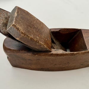 May include: A small, antique wooden block plane with a dark brown finish. The blade is partially exposed, and the plane has a curved base. This woodworking tool is designed for smoothing and shaping wood surfaces.