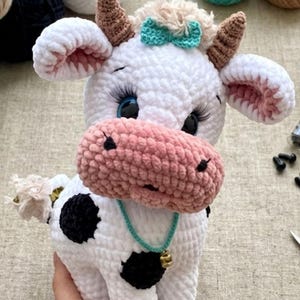 May include: A handmade crochet cow plushie. The white cow has black spots, a pink muzzle, and blue eyes with long eyelashes. It has brown horns, a teal bow, and a turquoise necklace with a gold bell. The cow is a soft, cuddly toy.