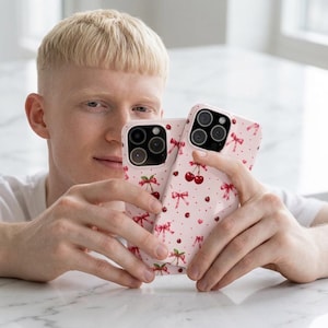 Puede incluir: Dos fundas para smartphone de color rosa claro con un patrón repetitivo de cerezas rojas, pequeños lazos rosas y diminutos puntos rojos. Cada funda tiene un recorte para un sistema de triple cámara en la parte trasera y acabado brillante.