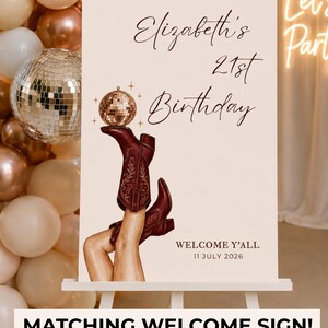 May include: A rectangular sign on a white easel featuring the text Elizabeth's 21st Birthday and Welcome Y'all 11 July 2026. The design includes an illustration of legs wearing dark red cowboy boots, with one boot balancing a small disco ball.