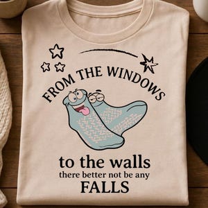 May include: A beige t-shirt with the text "From the windows to the walls there better not be any FALLS" and a cartoon image of two socks. The shirt is folded on a wooden surface.