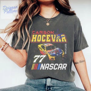 May include: Dark gray t-shirt featuring a NASCAR design. The shirt displays the name "CARSON HOCEVAR" above a colorful race car graphic with the number "77" and the word "NASCAR" below.