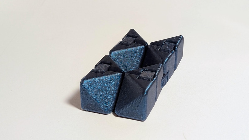 May include: A geometric puzzle toy in shades of dark blue. The puzzle is composed of several interlocking triangular pieces, each with a textured surface and small square details. The toy is designed to be manipulated and reconfigured.