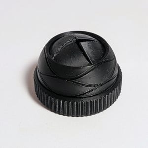 May include: A black, 3D-printed object resembling a camera lens aperture. The object has a circular base with ridged edges and a domed top with curved, overlapping segments. The aperture is partially open.