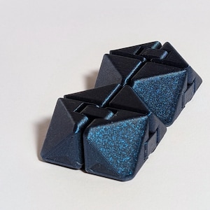May include: Two dark blue, geometric, interlocking cubes with a glittery, textured surface. The cubes are positioned on a white background. The design features sharp angles and a modern aesthetic.
