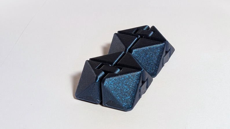 May include: Two dark blue, geometric, interlocking cubes with a glittery, textured surface. The cubes are positioned on a white background. The design features sharp angles and a modern aesthetic.