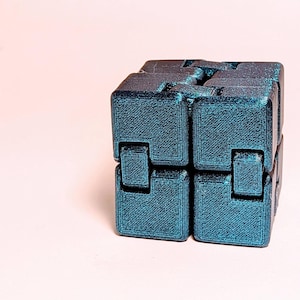 May include: A teal-colored infinity cube toy, composed of interconnected blocks. The cube is made of a textured material and sits on a light pink surface. The design allows for continuous folding and unfolding.