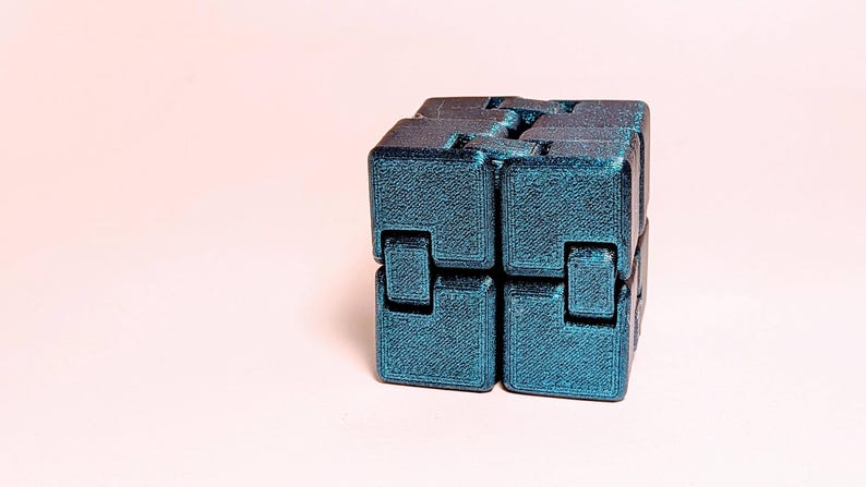 May include: A teal-colored infinity cube toy, composed of interconnected blocks. The cube is made of a textured material and sits on a light pink surface. The design allows for continuous folding and unfolding.