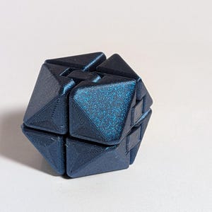 May include: A dark blue, geometric puzzle cube with a textured, glittery surface. The cube is composed of several interlocking triangular and rectangular pieces, creating a complex, three-dimensional shape. The puzzle is designed to be manipulated and solved.