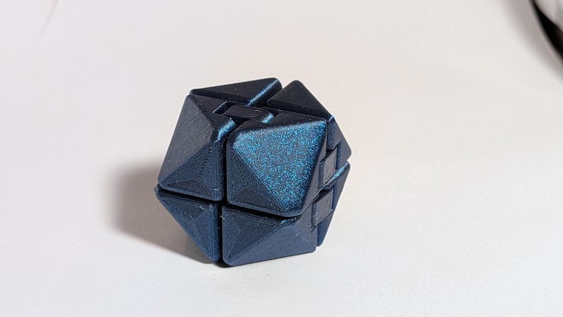 May include: A dark blue, geometric puzzle cube with a textured, glittery surface. The cube is composed of several interlocking triangular and rectangular pieces, creating a complex, three-dimensional shape. The puzzle is designed to be manipulated and solved.