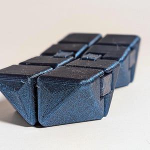 May include: A dark blue, articulated fidget toy, composed of several connected triangular blocks. The blocks are arranged in a zig-zag pattern, creating a flexible, tactile object. The toy is designed for stress relief and sensory stimulation.