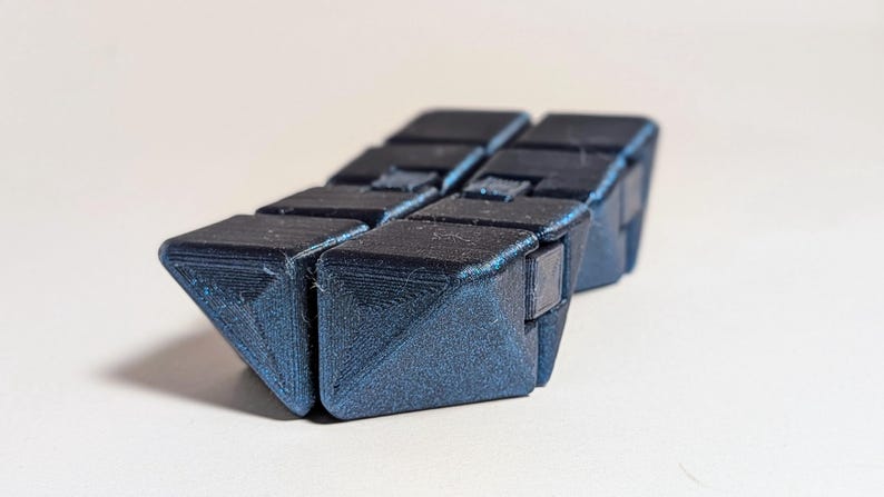 May include: A dark blue, articulated fidget toy, composed of several connected triangular blocks. The blocks are arranged in a zig-zag pattern, creating a flexible, tactile object. The toy is designed for stress relief and sensory stimulation.